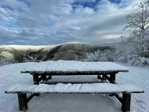 A snowy picnic table overlooks a winter landscape of mountains and trees under a cloudy sky.