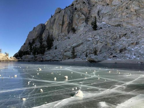 A frozen lake with ice patterns and small rocks, surrounded by rocky cliffs and trees under a clear blue sky.