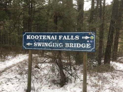 Signpost in a snowy forest directing to Kootenai Falls and Swinging Bridge, with arrows pointing in both directions.