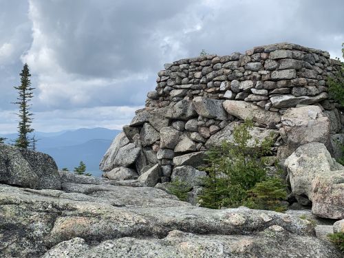 Stone structure atop a rocky mountain, surrounded by trees and distant hills under a cloudy sky.
