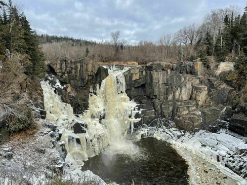 A scenic waterfall cascading over rocky cliffs, surrounded by trees and a partially frozen river below.