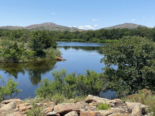 A serene landscape featuring a calm lake surrounded by greenery and rocky terrain, with mountains in the background.
