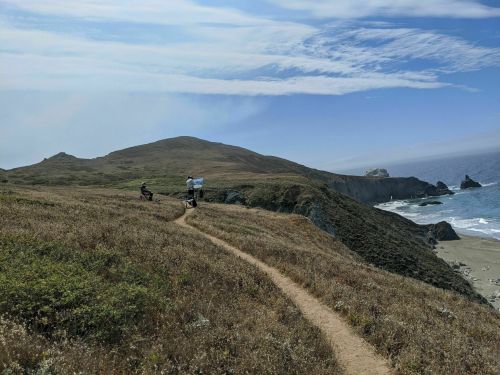 Two hikers on a trail overlooking a coastal landscape with cliffs and ocean under a blue sky with wispy clouds.