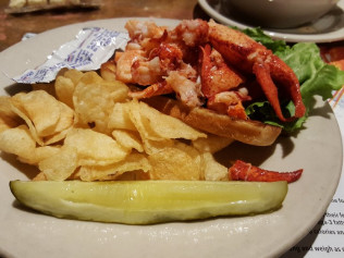 A lobster roll on a plate with lettuce, potato chips, and a pickle on the side.