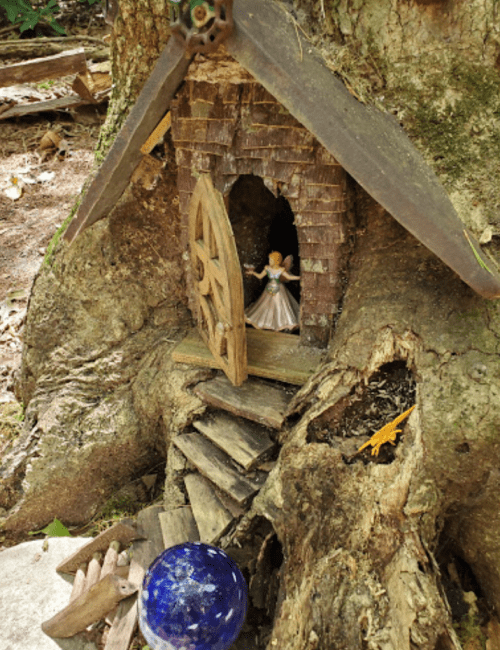 A whimsical fairy house built into a tree trunk, with a small figure and a blue glass orb nearby.