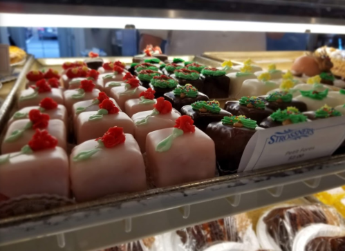 A display of colorful pastries, including small cakes topped with red roses and green leaves, in a bakery case.