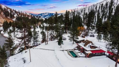 A snowy landscape featuring a cozy cabin surrounded by pine trees and mountains, with a small pool in the yard.
