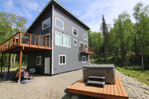 A modern gray house with multiple windows, a deck, and a hot tub, surrounded by trees and gravel.