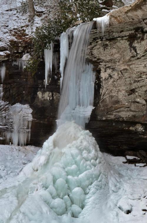 A frozen waterfall cascading over rocky cliffs, surrounded by ice formations and snow-covered ground.