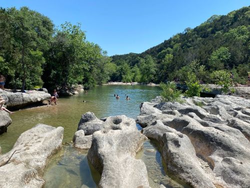A serene river scene with people swimming and relaxing among rocky banks and lush greenery under a clear blue sky.