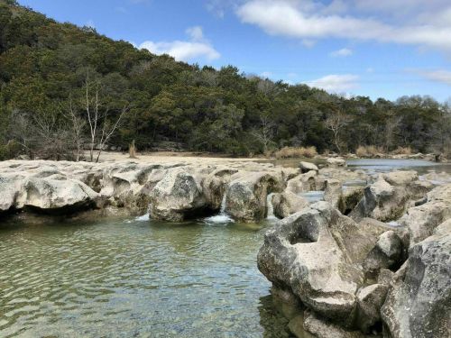 A serene landscape featuring rocky formations by a clear stream, surrounded by lush greenery and a blue sky.