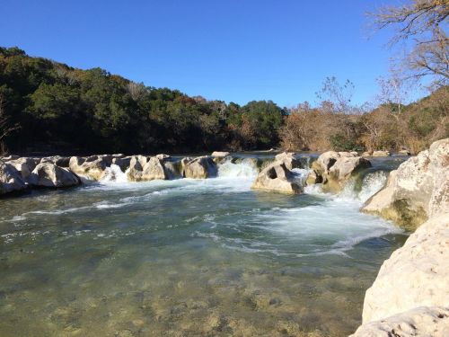 A clear river flows over rocky formations, surrounded by trees under a bright blue sky.