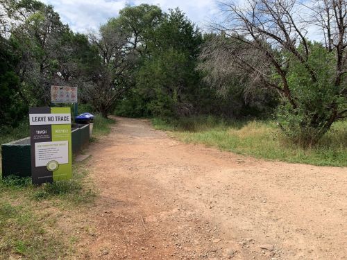 A dirt path leads into a wooded area, with a sign promoting "Leave No Trace" and recycling options nearby.