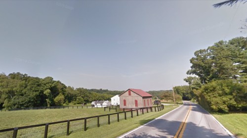 A rural scene featuring a road, a red barn, and green fields with trees in the background under a clear sky.