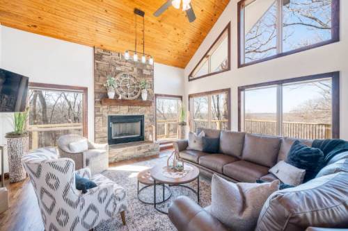 Cozy living room with a stone fireplace, large windows, and wooden ceiling, featuring comfortable seating and a coffee table.