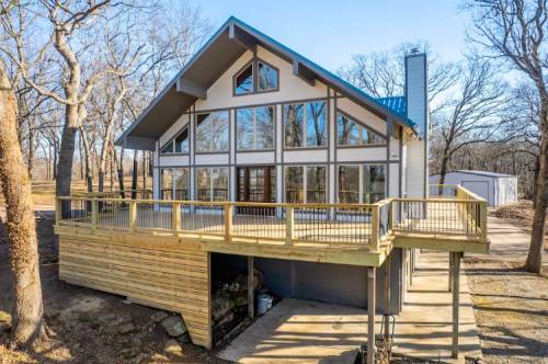 Modern house with large windows and a wooden deck, surrounded by trees and a clear blue sky.