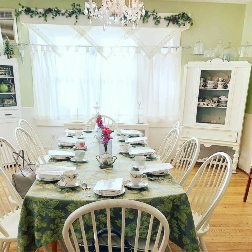 A bright dining room with a green tablecloth, set for tea, featuring white chairs and a chandelier above.