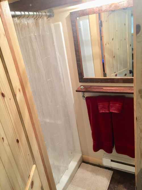 A small bathroom with a shower, mirror, wooden accents, and red towels hanging on a rack.