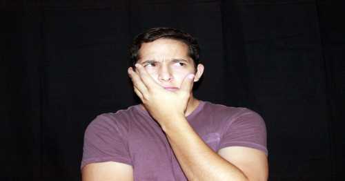 A young man with a puzzled expression rests his chin on his hand against a black background.