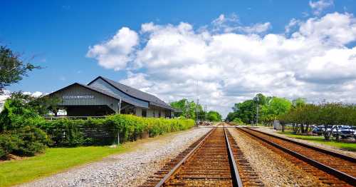 A view of a railway track leading to a quaint train station under a bright blue sky with fluffy clouds.