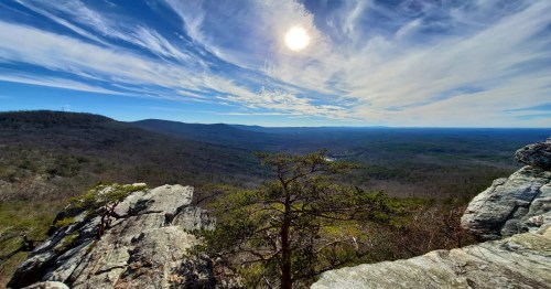 A panoramic view of mountains and valleys under a bright sun, with a tree in the foreground and wispy clouds in the sky.