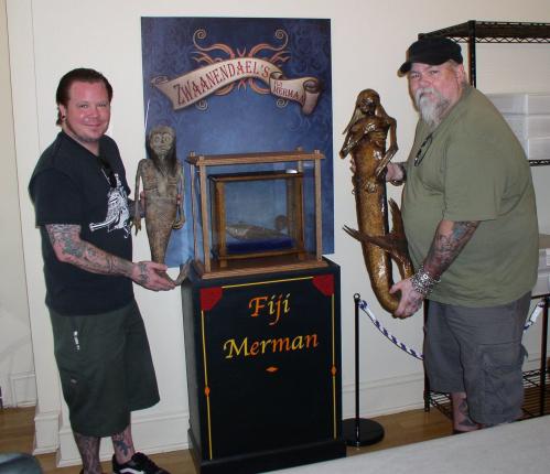 Two tattooed men hold artifacts next to a display labeled "Fiji Merman," featuring a glass case and statues.
