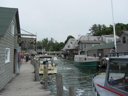 A quaint harbor scene with wooden buildings, boats docked, and people walking along a narrow path by the water.