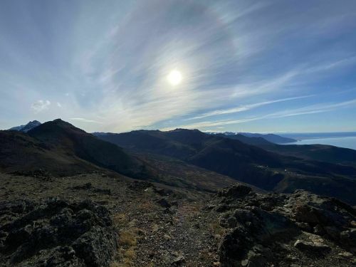 A panoramic view of mountains under a bright sun, with a clear sky and distant ocean visible on the horizon.