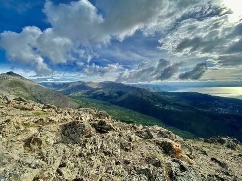 A panoramic view of rugged mountains and valleys under a dramatic sky with clouds, overlooking a distant coastline.