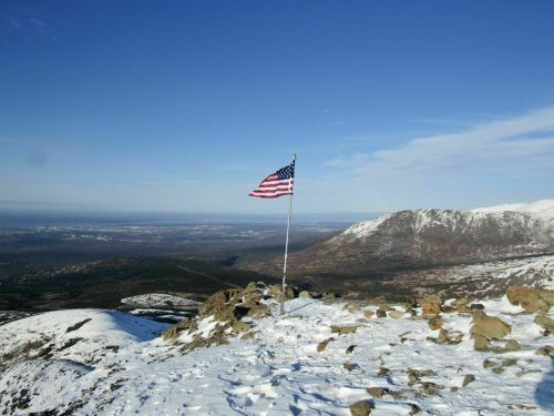 A snowy mountain landscape with an American flag waving on a pole against a clear blue sky.