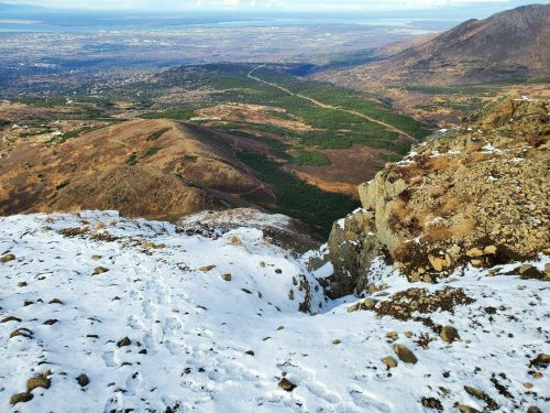 A panoramic view of a mountainous landscape with snow, rocky terrain, and a valley below, stretching into the distance.