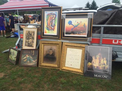 A collection of framed artworks and photographs displayed on the ground at a flea market, with a truck in the background.