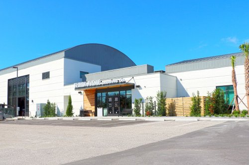 Modern building with a curved roof, large windows, and a welcoming entrance, surrounded by greenery and a parking lot.