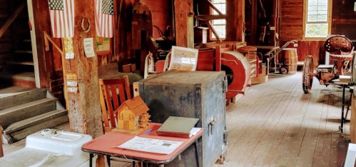 Interior of a rustic barn with wooden floors, vintage tools, a safe, and a model house on a table. American flags hang on the walls.