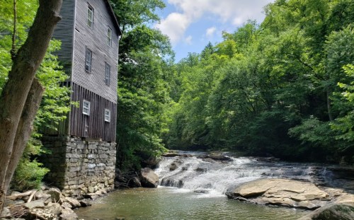 A historic mill stands beside a flowing creek, surrounded by lush green trees and rocky terrain under a blue sky.