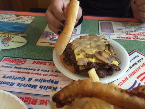 A hand holds a bun above a plate with a cheeseburger topped with onions and mushrooms, alongside curly fries.