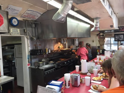 A busy diner kitchen with chefs cooking, stainless steel appliances, and patrons enjoying meals at red-topped tables.