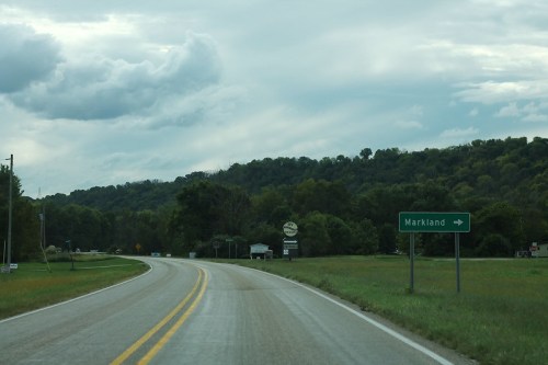 A winding road leads to a sign for Markland, surrounded by green hills and cloudy skies.