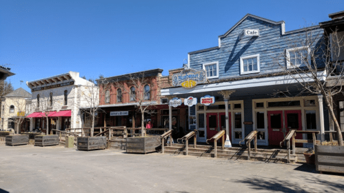 A row of colorful, Western-style storefronts with wooden walkways and clear blue skies above.