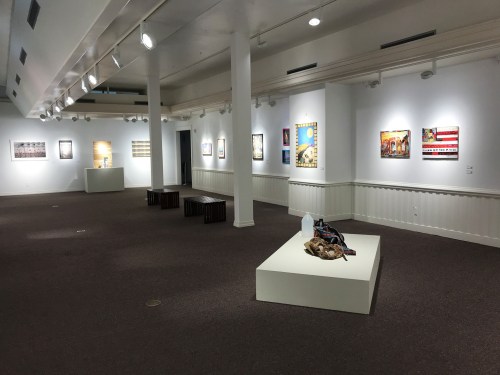 An art gallery with white walls, featuring various framed artworks and a central display on a white pedestal.