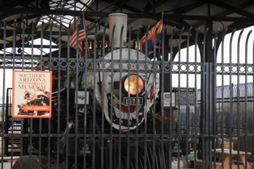 A vintage steam locomotive behind a black fence, with flags and a sign for the Southern Arizona Transportation Museum.