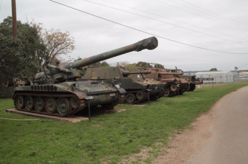A row of military tanks displayed outdoors along a gravel path, with trees and a building in the background.