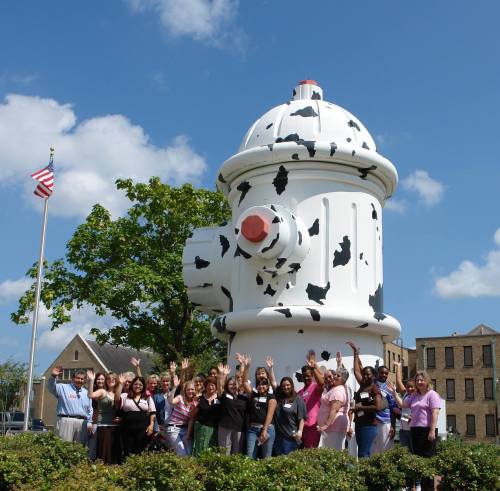 A group of people stands in front of a large, decorative fire hydrant, waving and smiling under a blue sky.