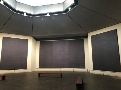 A minimalist art gallery featuring large, dark canvases on the walls and wooden benches in the center.