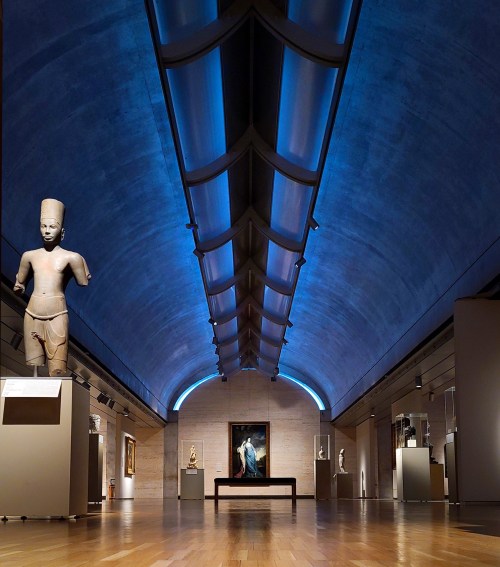 A spacious museum gallery featuring sculptures, paintings, and blue-lit architectural details.