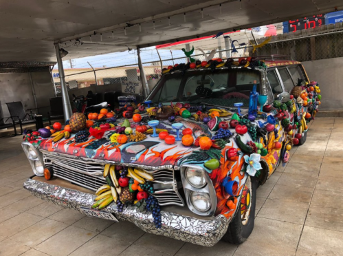 A colorful, fruit-decorated car covered in various artificial fruits and flowers, parked under a shelter.