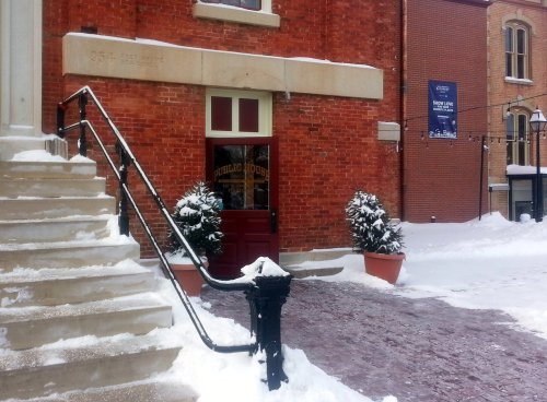 Entrance of a brick building with snow-covered steps and potted plants, surrounded by a winter landscape.