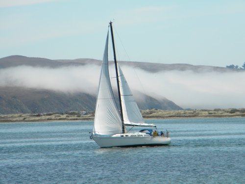 A sailboat with white sails glides across calm waters, surrounded by misty hills in the background.