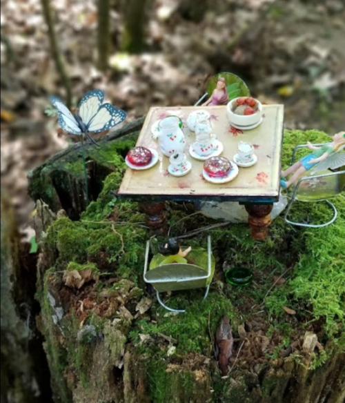 A whimsical miniature tea set on a mossy stump, with a butterfly nearby and tiny figures enjoying the scene.