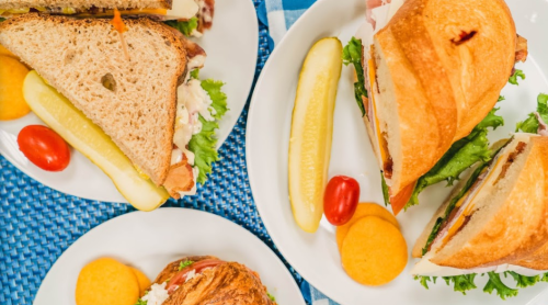 A top view of sandwiches on plates, accompanied by pickles, cherry tomatoes, and round crackers on a blue tablecloth.